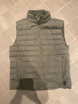 Grön quiltad dunväst med dragkedja från dressman  - Snygg grön dunväst med quiltad design och hög krage. Västen har två fickor med dragkedja och en diskret logotyp på ena fickan. Perfekt att slänga över en hoodie för extra värme. Stängs med dragkedja framtill.