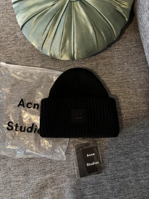 Svart stickad mössa från Acne Studios - Svart ribbstickad mössa från Acne Studios med klassisk smiley-logga framtill. Tillverkad i 100% ull för extra värme och komfort. Snygg och enkel design som passar till alla dina outfits. Kommer med originalpåse och tag.