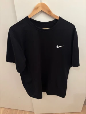 Svart Nike t-shirt med logga - Svart t-shirt från Nike med klassisk vit swoosh-logga på bröstet. Enkel och stilren design med rund hals och korta ärmar. Perfekt för dig som gillar sportiga och cleana plagg. Materialet känns mjukt och bekvämt mot huden.
