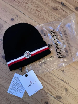 Svart Moncler mössa - Svart ribbstickad mössa från Moncler med klassisk logotyp framtill. Mössan har en bred kant med vita och röda ränder som ger en sportig vibe. Perfekt för kalla dagar och snygg till streetwear. Materialet är mjukt och värmande, troligen ull eller syntet. Köpt här på Plick.