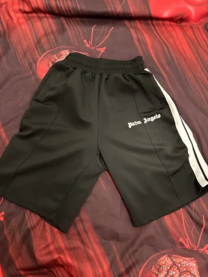 Palm Angels svarta shorts med vita ränder - Svarta shorts från Palm Angels med vita kontrasterande ränder längs sidorna och logotyp broderad på vänster ben. Modellen har elastisk midja och två sidofickor. Perfekta för en sportig och chill stil.