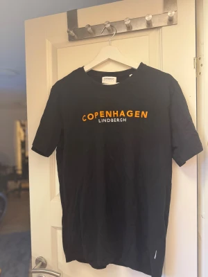 Svart t-shirt Lindbergh L - Säljer en svart t-shirt från Lindbergh med trycket 'Copenhagen' i orange och 'Lindbergh' i vitt på bröstet. Knappt använda i bra skick. 