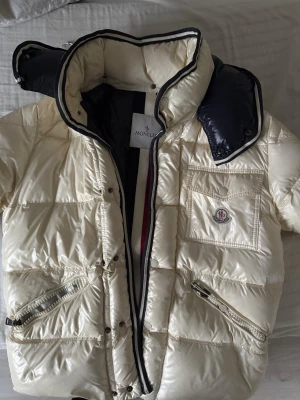 Vit Moncler dunjacka med huva - Säljer en snygg vit Moncler dunjacka med svart innerfoder och avtagbar huva. Jackan har dragkedjor på fickorna, bröstficka med Moncler-logga och coola randiga detaljer på insidan. Perfekt för kalla dagar när du vill hålla stilen.