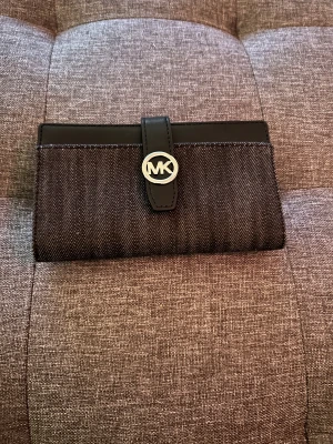 Svart plånbok från Michael Kors - Snygg svart plånbok från Michael Kors med MK-logga i silver på framsidan. Plånboken har flera kortfack, fönster för ID och dragkedja för mynt. Materialet är en mix av skinn och denim, vilket ger en cool och modern look.