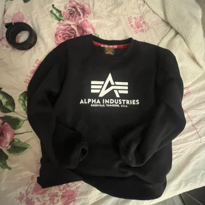 Svart Alpha Industries sweatshirt - Svart sweatshirt från Alpha Industries med stor vit logotyp och text på bröstet. Tröjan har rund hals och långa ärmar, insidan är mjuk och skön. Perfekt för dig som gillar streetwear och vill ha en enkel men snygg look.