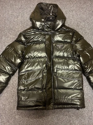 Svart pufferjacka från Prada - Säljer en svart, glansig pufferjacka från Prada med huva och stora fickor framtill. Jackan har klassisk quiltad design, dragkedja och ikonisk Prada-logga på bröstet. Perfekt för kalla dagar och riktigt snygg streetstil.