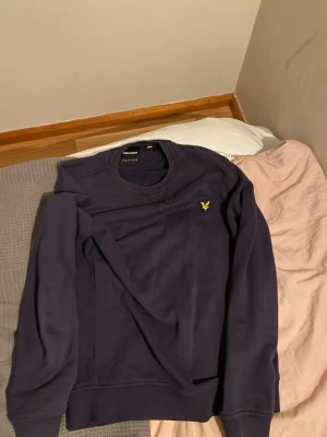 Mörkblå sweatshirt från Lyle & Scott - Mörkblå sweatshirt från Lyle & Scott med klassisk rund hals och gul broderad logga på bröstet. Tröjan har långa ärmar och ribbade muddar vid ärmslut och nederkant. Perfekt för en chill och stilren look.