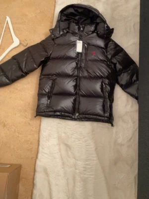 Svart pufferjacka från Polo Ralph Lauren - Säljer en svart pufferjacka från Polo Ralph Lauren med huva och flera praktiska fickor med dragkedja. Jackan har en glansig finish, elastiska muddar och RL-detaljer på dragkedjorna. Perfekt för kalla dagar och har en klassisk, sportig look.