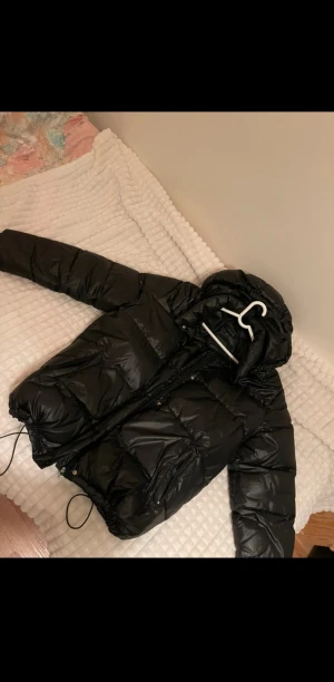 Svart pufferjacka  - Säljer en svart pufferjacka med huva som har glansig yta. Jag själv har inte storlek L men har använd den eftersom den kan vara ”baggy/oversized” om man vill det.  Perfekt för kalla dagar.