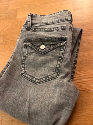 Grå bootcut jeans från Gina Tricot - Snygga grå jeans från Gina Tricot med bootcut-ben och klassisk femficksdesign. Jeansen har en silverfärgad knapp, bälteshällor och en cool tvättad look. Materialet är mjukt denim och passformen är normal. Perfekt för dig som gillar retrovibbar och stilren look.