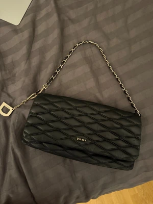Svart quiltad axelväska från DKNY - Snygg svart axelväska från DKNY med quiltat mönster och siljver kedja som axelrem. Väskan har en rektangulär form och DKNY-logga i metall framtill. Materialet är mjukt skinn och detaljerna ger en lyxig vibe. Perfekt storlek för det viktigaste och stilren look.