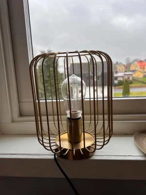 Snygg bordslampa i guld - modern design - äljer en stilren bordslampa i guldfärgad metall med dekorativ ”bur”-design. Lampan ger ett mysigt sken och passar perfekt i fönster, på nattduksbord eller som detalj i vardagsrummet. Fint skick och fungerar utan problem.  Mått (ca): – Höjd: ~25–30 cm – Bredd: ~18–20 cm  Ingår: – Lampfot – Glödlampa