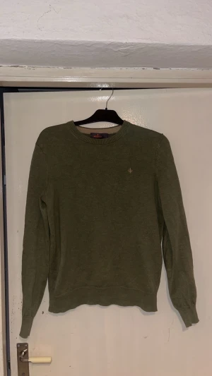 Olivgrön stickad tröja från Morris - Säljer en olivgrön stickad tröja från Morris med rund hals och diskret broderad logga på bröstet. Tröjan har ribbade muddar vid ärmslut och nederkant samt långärmad passform. Perfekt för lager-på-lager under kyliga dagar.