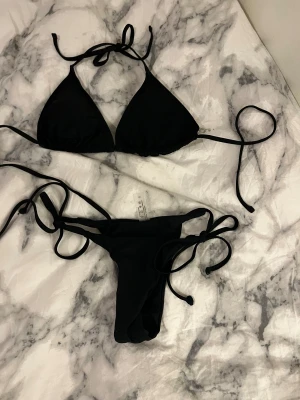 Svart bikini med knytband - Svart bikini med trekantsformad överdel och knytband både i nacken och ryggen. Underdelen har smala knytband på sidorna för justerbar passform. Från shein där av pris. Säljer för den ej passar, bara provad en gång över kläder. Överdel - L, Underdel - S