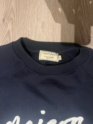 Mörkblå Maison Kitsuné sweatshirt - Snygg mörkblå sweatshirt från Maison Kitsuné med vit logotyp framtill. Tröjan har rund hals, långa ärmar och är gjord i mjuk bomull. Perfekt för dig som gillar stilrena och trendiga plagg med streetkänsla.