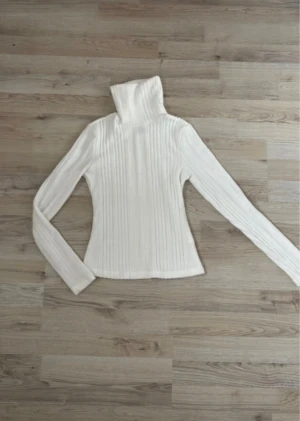 Vit ribbad polotröja-långärmad  - Klassisk vit polotröja i mjukt ribbstickat material. Stretchig och bekväm med hög, vikbar polokrage. Sitter snyggt och figurnära utan att bli obekväm. Perfekt basplagg till höst och vinter som värmer