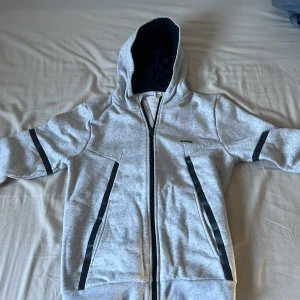 Grå tjocktröja med zip och luva  från HUGO BOSS  - Snygg grå tjocktröja från BOSS med svarta ränder längs ärmar och fickor. Tröjan har hög krage, hel dragkedja och en clean look med diskret BOSS-logga på bröstet. Perfekt för dig som gillar stilren och sportig vibe.