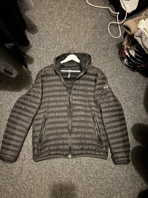 Mörkgrå pufferjacka från Colmar - Säljer en mörkgrå pufferjacka från Colmar med huva och två sidofickor med dragkedja. Jackan har ett quiltat mönster och Colmar-logga på vänster ärm. Perfekt för dig som gillar stilren och sportig look. Passar dig som vill ha en jacka med normal passform och lång ärm.