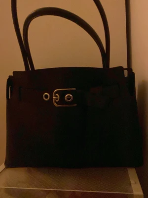 Svart axelväska med remdetalj från H&M - Jag säljer en axelväska från h&m! Det är lite en intressekoll om någon hade velat köpa men också att jag gärna vill få sålt den då jag ska köpa en annan väska mer i min stil🥰 Jag har använt den väldigt mycket men har inga stora defekter förutom att den kan vara lite smutsig, men inget man tänker på! Banden är typ lite ”böjda” så de sticker fram lite men inget enligt mig som stör eller ser konstigt ut☺️ Osäker på pris så man får komma med bud och man får såklart fråga frågor eller fråga om bilder🫰🏻