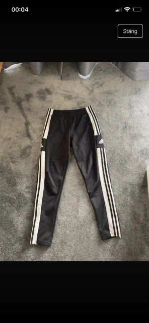 Svarta Adidas träningsbyxor med vita ränder - Svarta träningsbyxor från Adidas med klassiska vita ränder längs benen och logga på sidan. Byxorna har elastisk midja och smal passform, perfekta för sport eller chill. Materialet är syntet och de har en modern, sportig look.