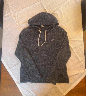 Blågrå hoodie från Hollister - Snygg mörkgrå hoodie från Hollister i storlek S. Tröjan har vit snörning i huvan, lång ärm och ett broderat Hollister-logo på bröstet. Materialet är mjukt och känns som bomull, perfekt för chill dagar. Passformen är normal och färgen är melerad mörkgrå.