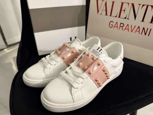 Valentino Rockstud sneakers vit/rosa - Snygga Valentino Rockstud sneakers i vitt skinn med ljusrosa band över foten dekorerat med ikoniska pyramidnitar. Rund tå, platt sula och snörning framtill. Perfekt för dig som gillar lyxiga detaljer och streetstyle.