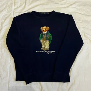 Mörkblå Polo Bear tröja Ralph Lauren - Snygg mörkblå sweatshirt från Polo Ralph Lauren med ikoniskt Polo Bear-tryck på bröstet. Tröjan har rund hals, långa ärmar och ribbade muddar. Perfekt för dig som gillar klassisk streetstyle med en twist. Materialet är mjuk bomull för skön komfort.