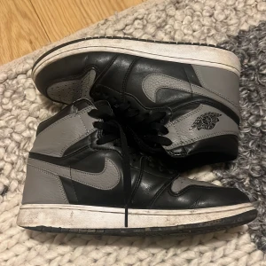 Nike Air Jordan 1 svart/grå high - Nike Air Jordan 1 high sneakers i svart och grått skinn med klassisk Jordan-logga på sidan. Skorna har snörning, perforerad tå och vit platt sula. Snygg kontrast mellan de mörka och ljusa panelerna, perfekt för dig som gillar streetwear och retrostil.