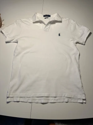 Vit piké från Polo Ralph Lauren - Vit klassisk pikétröja från Polo Ralph Lauren i slim fit-modell. Tröjan har korta ärmar, krage med två knappar och den ikoniska blå broderade loggan på bröstet. Tillverkad i mjuk bomull som är skön mot huden och passar perfekt till jeans eller shorts.