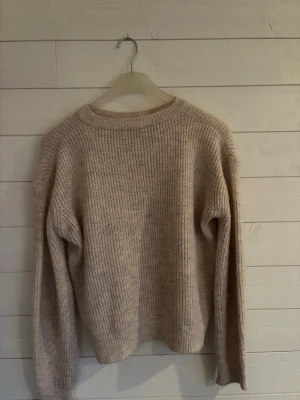 Beige stickad tröja med rund hals - Mysig beige stickad tröja med rundad hals och ribbade muddar. Tröjan har en loose passform och långa ärmar, perfekt för att hålla värmen under kyliga dagar. Enkel och stilren design som funkar till många olika outfits. Storleken är antingen XS eller S