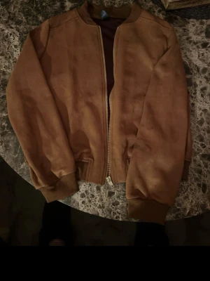 Brun mocka bomberjacka med dragkedja - Snygg brun bomberjacka i mocka med dragkedja framtill och ribbade muddar vid ärmslut och midja. Jackan har en klassisk, stilren look och passar perfekt för dig som vill ha en tidlös stil med edge.