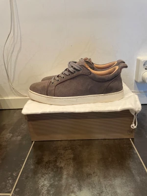 Grå mocka sneakers från Louboutin - Snygga grå sneakers från Christian Louboutin i mjuk mocka med rund tå och platt Röd sula. Skorna har grå snörning och beige innersida, vilket ger en clean och lyxig vibe. Perfekta för dig som gillar stilrena och exklusiva sneakers.Priset är inte hugget i sten🪨 