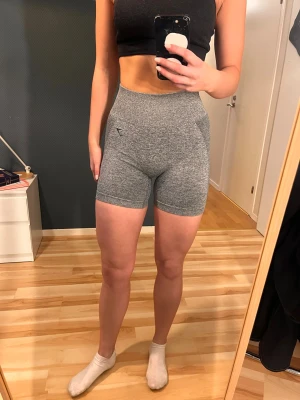 Grå höga träningsshorts från Gymshark - Säljer ett par helt oanvända, grå, högmidjade träningsshorts från Gymshark med svart logga på linningen. De är tighta och har en snygg, sportig passform som sitter skönt mot kroppen. Materialet är stretchigt och andas, perfekt för gymmet eller löprundan.