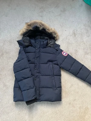Canada Goose Jacka - Säljer en mörkblå dunjacka från Canada Goose med fluffig päls på huvan och klassisk logga på ärmen. Perfekt för kalla vinterdagar! Hör av dig vid minsta fråga! Pris ej hugget i sten!