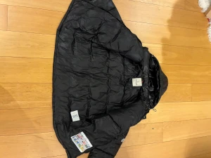 Svart dunjacka från Moncler - Svart dunjacka från Moncler med klassisk quiltad design och dragkedja framtill. Jackan har en snygg glansig finish, huva och coolt Moncler-märke på ärmen. Insidan har en unik serietidningspatch och etikett med DIST-certifiering.