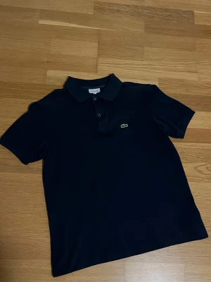 Lacoste Pike - Snygg mörkblå pikétröja från Lacoste med klassisk krage och två knappar framtill. Tröjan har det ikoniska gröna krokodilmärket broderat på bröstet. Materialet är bomull och modellen är kortärmad med normal passform.