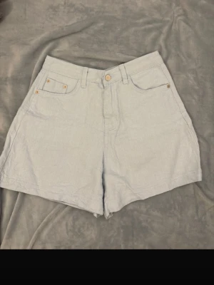 Ljusblåa högmidjade shorts i bomull - Storlek M - Snygga ljusblåa shorts med hög midja och klassisk femficksdesign. De har bälteshällor, knapp och dragkedja framtill samt två stora bakfickor. Materialet är mjukt bomullstyg som ger en chill och fräsch vibe, perfekt för varma dagar. 