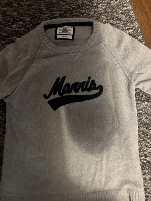 Grå stickad tröja Morris Gents M - Snygg grå stickad tröja från Morris Gents i storlek medium. Tröjan har svart Morris-tryck på bröstet och svarta armbågslappar på ärmarna. Rund hals med ribbad kant och ribbade muddar vid nederkant och ärmslut. Perfekt för dig som gillar klassisk stil med en twist.