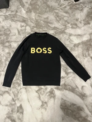 Svart BOSS sweatshirt med guldtryck - Svart sweatshirt från BOSS med stort guldigt BOSS-tryck på bröstet. Tröjan har rund hals, långa ärmar och ribbade muddar vid ärmslut och nederkant. Tillverkad i mjuk bomull som är skön att ha på sig. Perfekt för dig som gillar stilrena och snygga logotröjor. Kan även byta