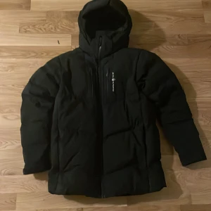 Svart vinterjacka från sail racing - Svart pufferjacka från sailracing med huva och dragkedja framtill. Jackan har en clean look med diskret logga på bröstet och är fodrad för extra värme. Perfekt för kalla dagar och har långa ärmar samt praktiska fickor. Snygg och funktionell design.