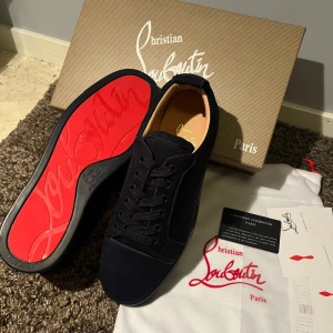 Mörkblå sneakers från Christian Louboutin - Snygga mörkblå sneakers från Christian Louboutin med lyxig mocka och klassisk röd sula. Skorna har rund tå, platt sula och snörning framtill. Diskret broderad logga på hälen och beige innersida. Perfekt för dig som gillar exklusiv stil och detaljer.
