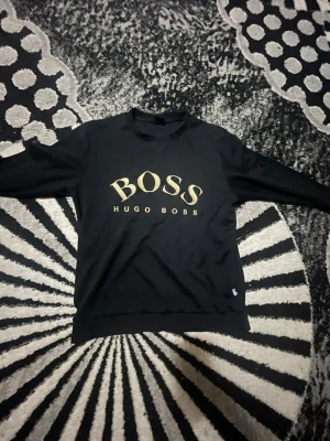 Svart sweatshirt från Hugo Boss - Svart sweatshirt från Hugo Boss med stort guldigt BOSS-tryck på bröstet. Tröjan har rund hals, långa ärmar och ribbade muddar vid ärmslut och nederkant. Materialet känns mjukt och bekvämt, perfekt för en chill och stilren look.