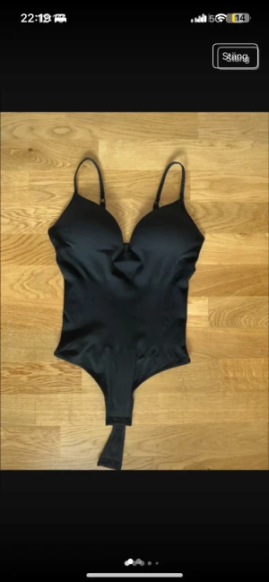 NY Svart bodysuit med smala axelband - Säljer en svart bodysuit med smala justerbara axelband och v-ringad form. Bodyn har en tight passform och knäppning i grenen. Materialet är mjukt och stretchigt, perfekt för att styla med jeans eller kjol. Enkel och stilren design utan mönster eller extra detaljer. Storlek M men passar S också. Helt ny, bara testad