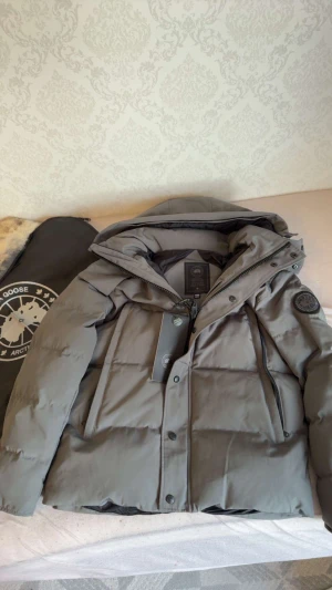 Grå dunjacka från Canada Goose - Säljer en grå dunjacka från Canada Goose med svart logotyp på ärmen. Jackan har huva, dragkedja och knappar framtill samt flera fickor. Materialet är slitstark polyester och dunfyllning, perfekt för kalla dagar. Medföljer en svart förvaringspåse med pälsdetalj. Storlek M