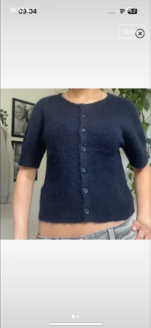 Mörkblå stickad kofta med kort ärm - Säljer en mörkblå stickad kofta med rund hals och korta ärmar. Koftan har en croppad passform och knäpps med sju matchande knappar framtill. Mjuk och fluffig känsla, perfekt för lager-på-lager. Passar dig som gillar enkel och stilren look.