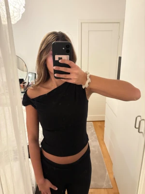 Svart offshoulder topp från H&M DIVIDED - Svart offshoulder topp från H&M DIVIDED i mjukt stretchigt material. Toppen har en snygg asymmetrisk axel med dragsko och knyt på ena sidan för en cool look. Perfekt att styla med jeans eller kjol för en trendig vibe.