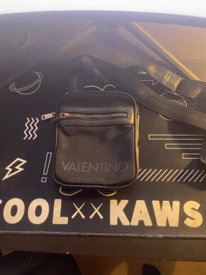 Svart axelväska från Valentino - Snygg svart axelväska från Valentino med justerbart axelband och dragkedja framtill. Väskan har ett stilrent tryck med logga och flera fack, både invändigt och utvändigt. Perfekt storlek för att bära med sig det viktigaste och passar till många olika outfits.