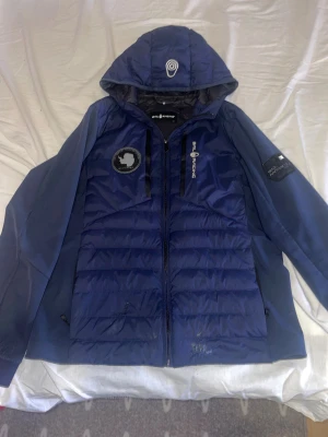 Mörkblå Sail Racing pufferjacka - Snygg mörkblå pufferjacka från Sail Racing med huva och coola patchar på bröstet och ärmen. Jackan har dragkedja framtill och quiltad design. Perfekt för dig som gillar sportig stil och vill ha något som sticker ut. finns några hål som har blivit fixade