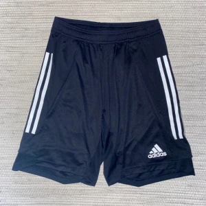Svarta Adidas shorts  - Svarta sportiga shorts från Adidas med klassiska vita ränder längs sidorna och Adidas-logga framtill. Modellen har elastisk midja och är gjorda i lätt polyester som andas. Perfekta för träning eller fotboll