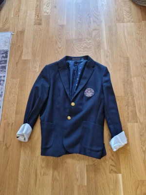 Mörkblå kavaj från GANT, strl 42 - Snygg mörkblå kavaj från GANT med guldfärgade knappar och broderad patch på bröstet. Fodret har ett diskret mönster och insidan är lyxigt satinliknande. Klassisk krage och två stora fickor framtill. Perfekt för dig som vill ha en stilren look. Använd fåtal gånger som ny